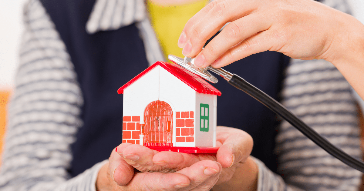 A senior woman holds a house in her hand as someone holds a stethoscope to it, symbolizing home care.